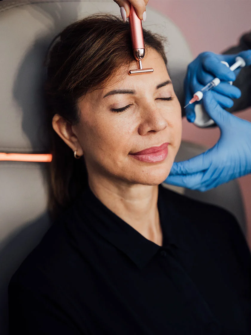 A woman receives a beauty treatment with a rose-gold microcurrent device on her forehead. A syringe is visible in the medical professional's other hand. The setting has soft lighting and a light beige background. - Botox and Wrinkle Relaxers in Miami Beach, FL