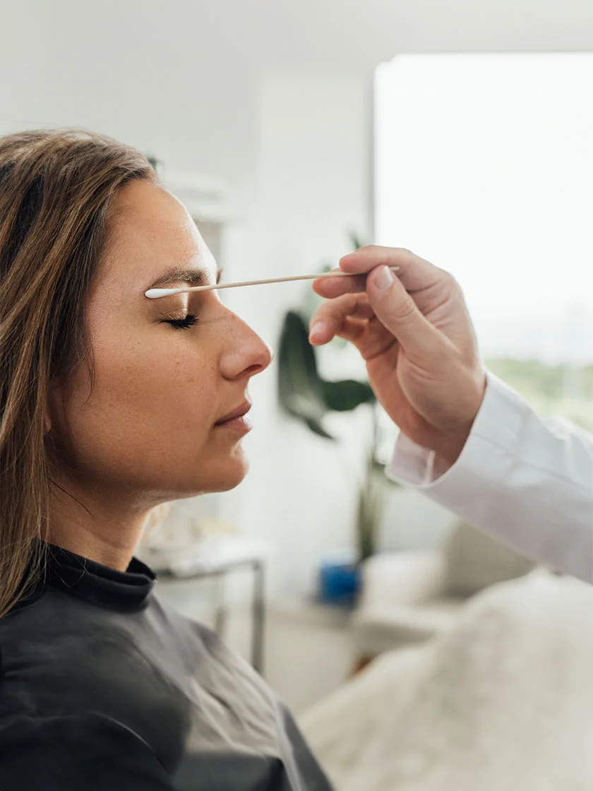 Close-up of a woman with shoulder-length brown hair receiving a cosmetic procedure. She has her eyes closed, and a medical professional in a white lab coat holds a cotton swab near her forehead. The blurred background suggests a clinic setting. - Browlift Forehead Lift in Miami Beach, FL