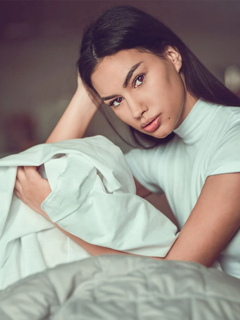 A young woman sits in bed, wrapped in a white sheet. Her dark hair cascades down her back as she looks directly at the camera with a neutral expression. Her makeup is subtle, and her smooth skin is visible. Her arms rest on the sheet, which is slightly wrinkled around her torso. The bedding beneath her is a light grayish-taupe color with a subtle pattern, and the background features muted, neutral tones. - Buccal Fat Removal in Miami Beach, FL