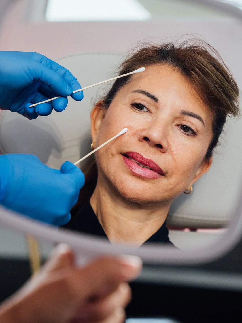 Close-up of a middle-aged woman looking into a mirror during a medical procedure. Cotton swabs are visible on her skin, with a person in blue gloves handling them. The background shows a medical facility. - Eyelid Surgery in Miami Beach, FL