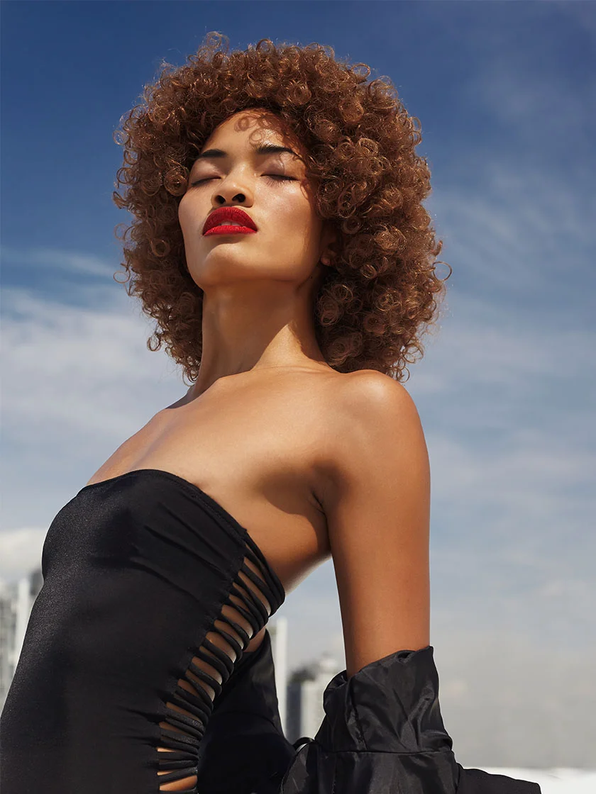 A woman with curly brown hair wears a black strapless swimsuit, her expression serene with closed eyes and red lipstick. The background shows a blue sky and blurred buildings, with natural light highlighting her confident posture. - Facial Balancing in Miami Beach, FL