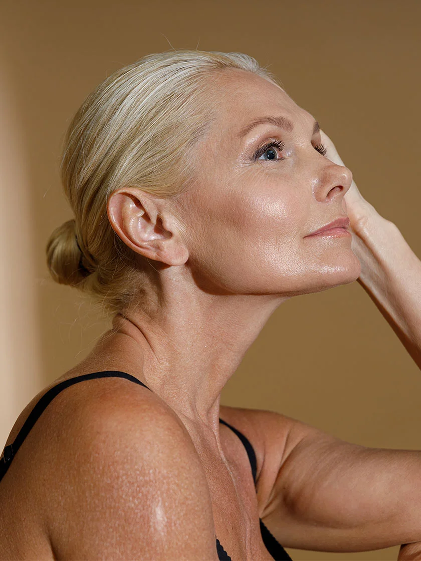 Close-up of a mature woman with gray hair in a bun, gazing upward with a neutral expression. Her skin shows wrinkles and fine lines, illuminated by soft lighting. She wears a black sleeveless top against a muted beige background. - Migrane Surgery in Miami Beach, FL